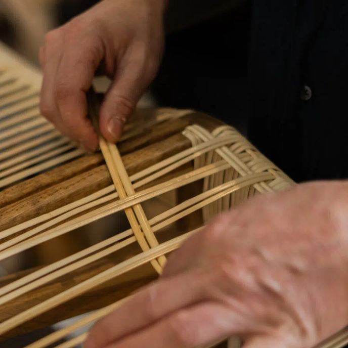 Hænder i gang med traditionel omfletning af sæde med lyst flet på mørk træramme.