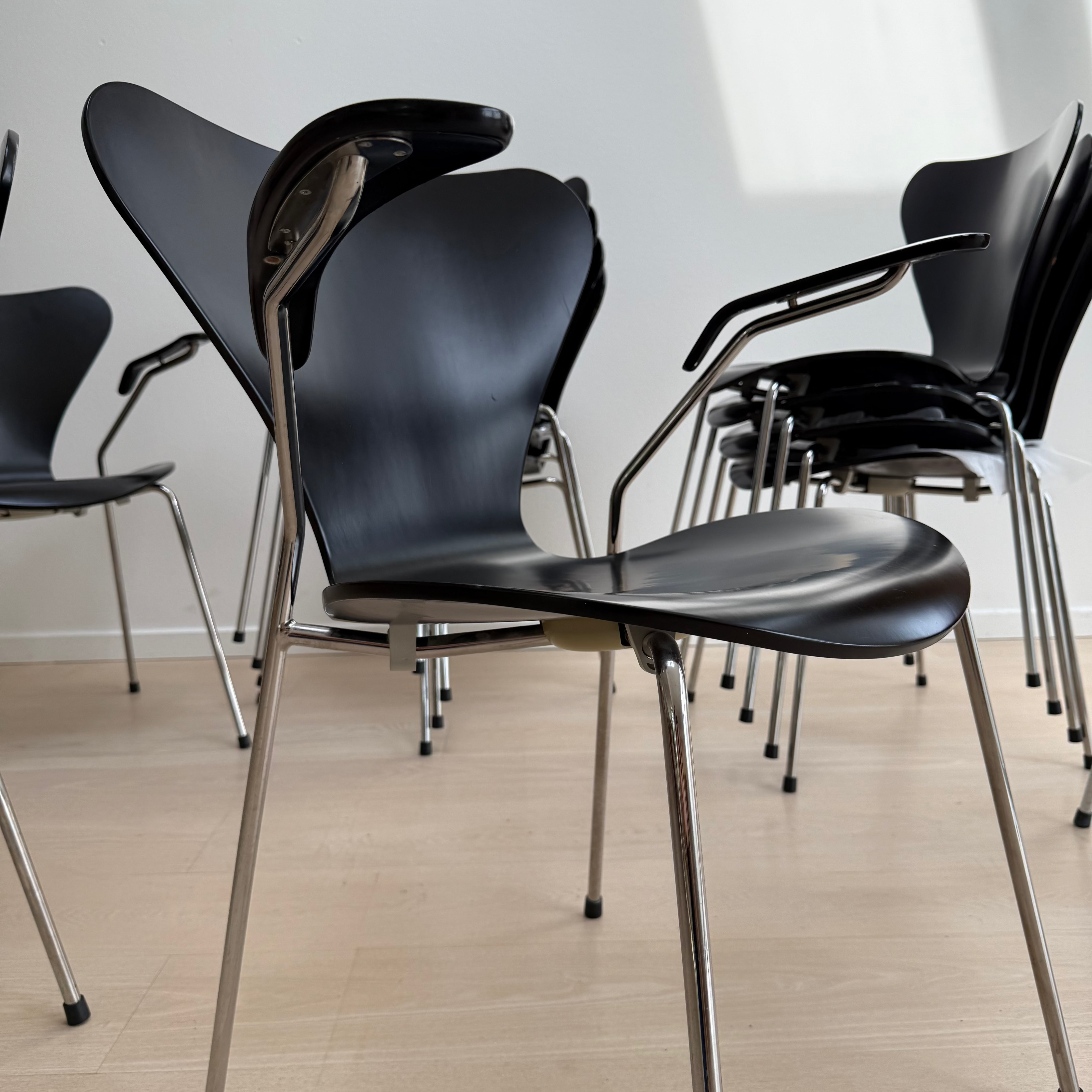 Sortlakerede Arne Jacobsen Syverstole / 7'er stole (model 3207) med armlæn og forkromede ben, opstillet i række på lyst trægulv.